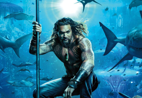 Aquaman berdiri tegak dengan trisula di tangannya, dikelilingi oleh berbagai jenis ikan hiu di bawah laut, menampilkan kekuatannya sebagai Raja Atlantis.