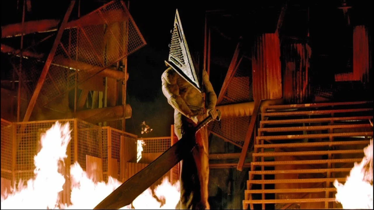 Pyramid Head berdiri menakutkan di tengah kebakaran dalam adegan dari film Return to Silent Hill, memegang pedang besar di tangan.