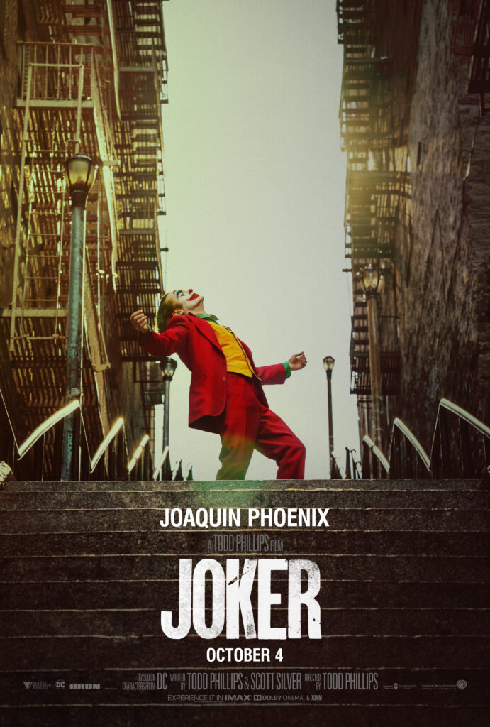 Joaquin Phoenix sebagai Joker berdiri di tangga kota, mengenakan jas merah ikonik dengan ekspresi penuh emosi, dari film Joker (2019).