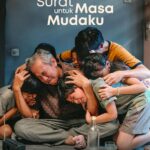 Poster film Surat untuk Masa Mudaku di Netflix yang menampilkan seorang ayah dan anak-anaknya saling berpelukan dengan ekspresi sedih di dalam rumah.