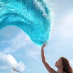 Seorang gadis mengangkat tangan menyentuh gelombang laut berbentuk lengkungan di langit dengan latar awan cerah, menggambarkan adegan dari Moana (Live-Action).