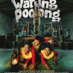 Poster film horor komedi Indonesia berjudul 'Warung Pocong' yang menampilkan tiga pria ketakutan bersembunyi di bawah meja kayu, sementara beberapa sosok hantu pocong berdiri di belakang mereka di dalam sebuah gudang atau warung tua yang remang-remang.
